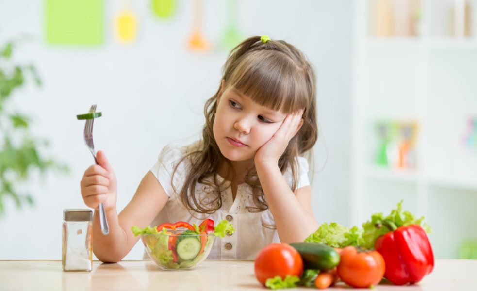 A girl toddler looks at a piece of vegetable on her fork, refuses to eat it