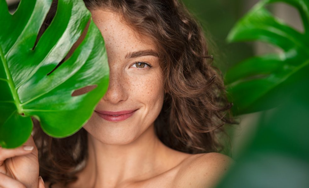 beautiful curly haired woman with freckles smiling behind huge leaves
